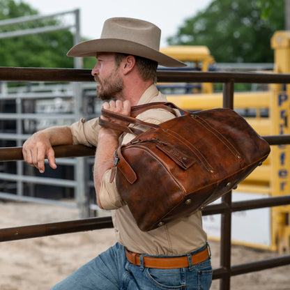 Lifestyle image of Cowboy leaning against fencing, holding leather travel duffle bag, see merchant for exact details