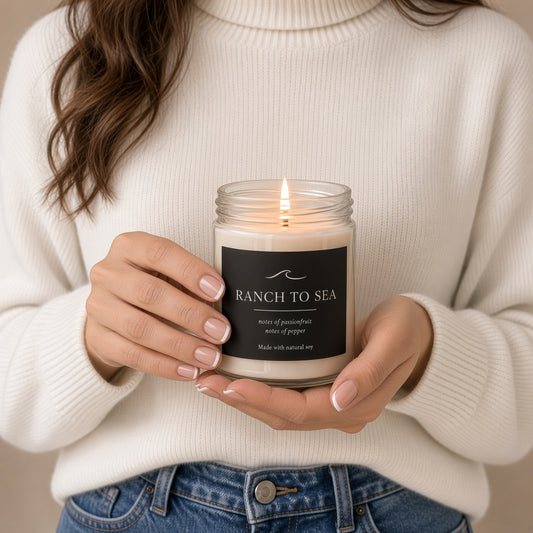 A person in a cream knit sweater holds a lit glass jar candle labeled 'RANCH TO SEA' with a black label and white wax. The person is also wearing blue jeans, and the background is a soft beige indoor setting.